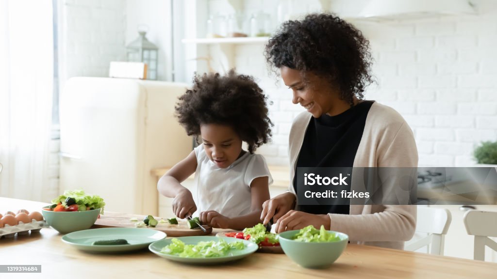 Happy biracial mom and little daughter cook together Smiling loving African American mother and little daughter prepare healthy delicious salad in kitchen. Happy caring biracial mom and small ethnic girl child cook together at home. Family concept. Healthy Eating Stock Photo