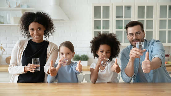 Portrait of young multiracial family with two children drink clean still mineral water together in kitchen. Smiling multiethnic mom and dad with diverse daughters show thumbs up recommend hydration.