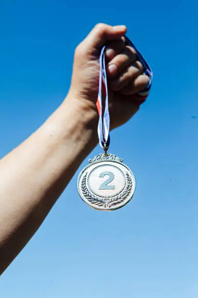 Photo of A hand holds up a second-place silver medal, with the sky in the background. Victory concept