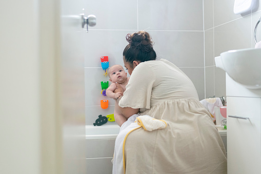 A young mother lifts her baby daughter out of the bath tub. She is getting ready to dry her off and get her to bed for the evening. In the background on the wall tiles, children's toys can be seen stuck onto them. White bathroom setting. Tender moment between mother and daughter.