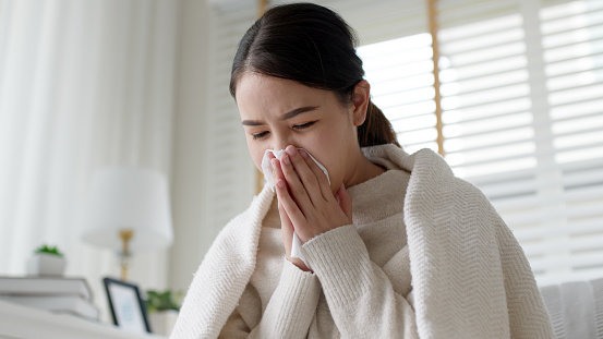 Sick young asian woman sitting under the blanket on sofa and sneeze with tissue paper at home. Female blowing nose, coughing or sneezing in tissue at home, suffering from flu. Cold and fever concept
