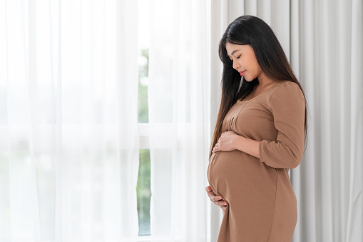 Asian happy pregnant woman is standing beside windows and touching her belly. pregnancy, motherhood, people and expectation concept