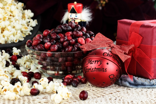 String of popcorn and cranberries with bowl of cranberries, popcorn, gift and ornaments in background. Shallow depth of field. 
