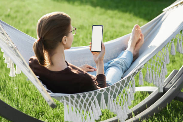 istock Woman relaxing in a hammock using smartphone with white blank empty screen mock-up 1319408945