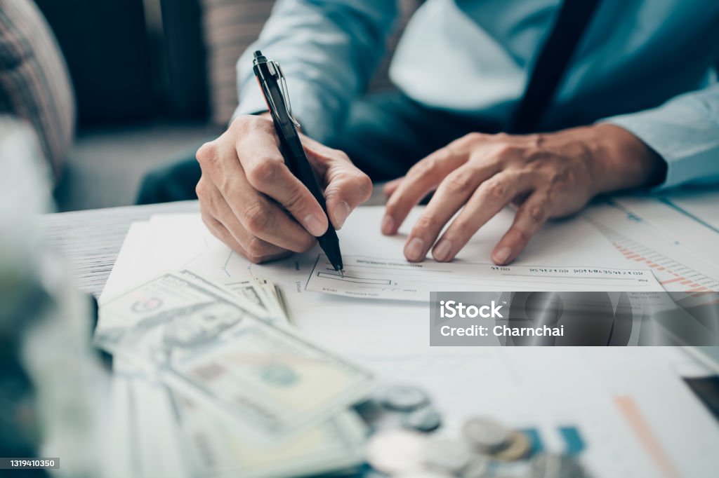 Business man hand writing and signing white blank bank check book and dollar bill, coin, laptop and graph chart on the desk at office. Payment by check, paycheck, payroll concept. Check - Financial Item Stock Photo
