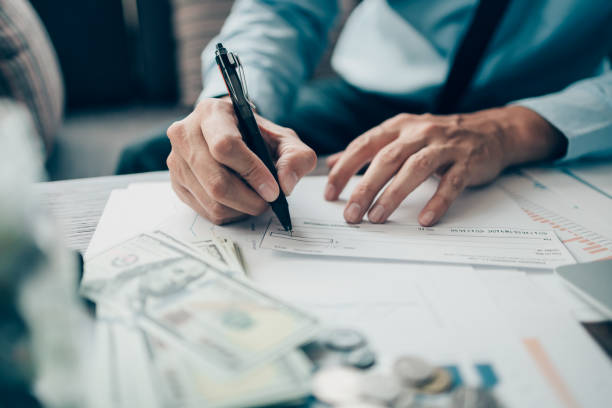 Business man hand writing and signing white blank bank check book and dollar bill, coin, laptop and graph chart on the desk at office. Payment by check, paycheck, payroll concept. stock photo