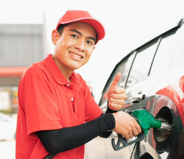 Asian gas station worker man give thumbs up in one hand and holding green fuel nozzle into one hand for filling high energy power fuel into black auto car tank in petrol station, commercial service for benzine, diesel, gasohol, gasoline. el hombre trabajador de la gasolinera asiática da pulgares hacia arriba en una mano y sosteniendo la boquilla de combustible verde en una mano para llenar el combustible de alta energía en el tanque de automóvil negro en la estación de servicio, servic