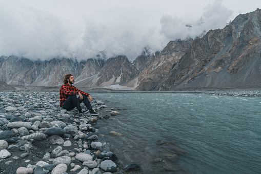 Young Caucasian man sitting  above the river on the background of  Karakoram mountain range in Pakistan