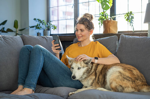 Mature woman relaxing on sofa with her dog at home using smart phone.