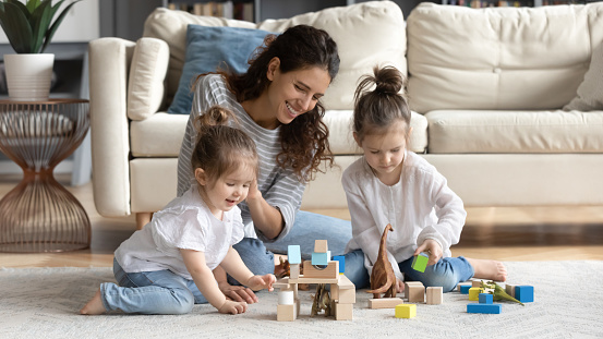 Happy caring young Caucasian mother have fun play with small daughter at home in living room. Smiling loving mom feel playful engaged in game activity with blocks bricks with little girls children.
