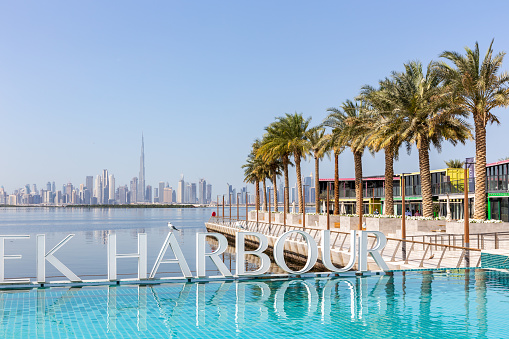 Dubai, UAE, 22.02.2021. Dubai Creek Harbour sign by turquoise water pool with rows of palm trees and Dubai Downtown skyline in the background.