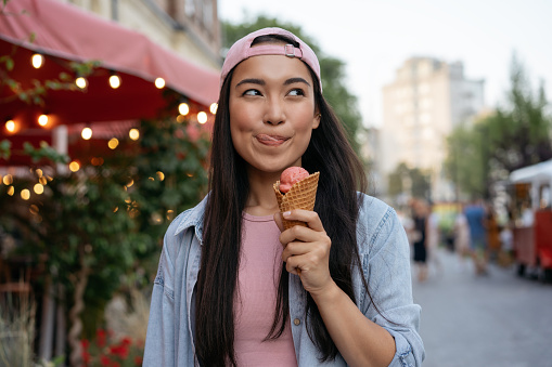 Hermosa mujer asiática comiendo helado en la calle. Hipster emocional con ropa casual sosteniendo sabroso postre de verano mirando hacia otro lado al aire libre. Festival gastronómico photo