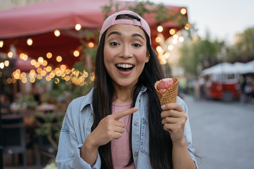Young excited asian woman eating ice cream on the street. Emotional hipster wearing casual clothing holding tasty dessert, selective focus. Food fair concept