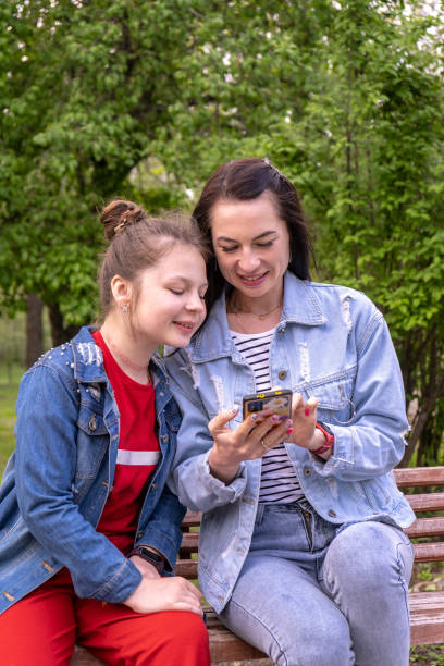 mother and teenage daughter walking in a park, sitting on a bench and looking in smartphone, happy young caucasian woman with long hair and teenage girl surfing the internet outdoors, lifestyle family - surfing wireless vertical outdoors lifestyles imagens e fotografias de stock