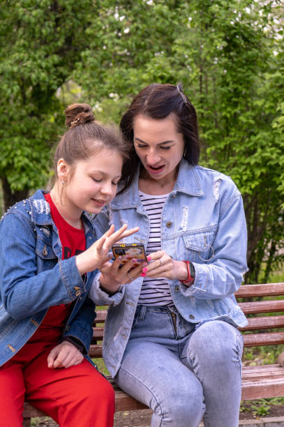 mother and teenage daughter walking in a park, sitting on a bench and looking in smartphone, happy young caucasian woman with long hair and teenage girl surfing the internet outdoors, lifestyle family - surfing wireless vertical outdoors lifestyles imagens e fotografias de stock