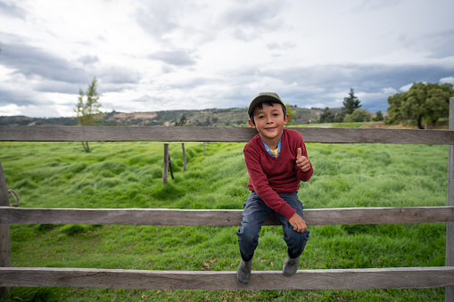 Portrait of a Happy Latin American boy sitting on a fence at a farm with thumbs up and looking at the camera smiling - lifestyle concepts