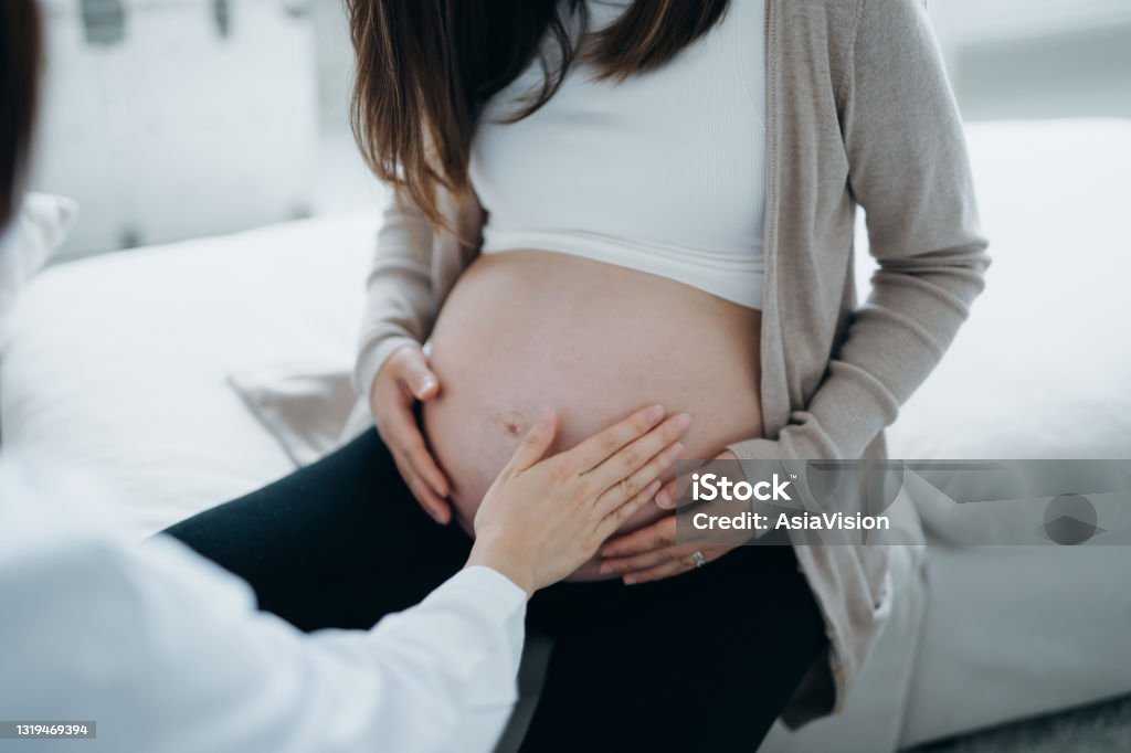 Cropped shot of Asian pregnant woman having a consultation with doctor during routine check up at clinic. Doctor is performing an examination and touching the belly. Check-ups, tests and scans to ensure a healthy pregnancy for both mother and unborn baby Midwife Stock Photo