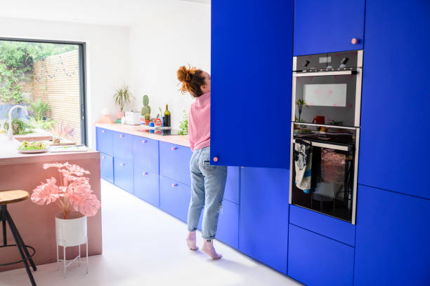 Mid adult Caucasian woman reaching into refrigerator Full length view of woman in jeans and casual top standing on tiptoe to access high shelf in vibrant blue kitchen. oven door stock pictures, royalty-free photos & images