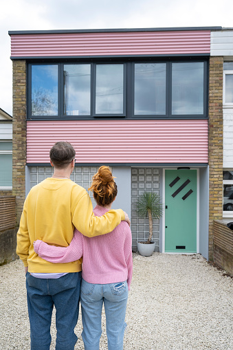 Rear view of man and woman in casual clothing standing outdoors with arms around each other as they look at their modern style two-story house.