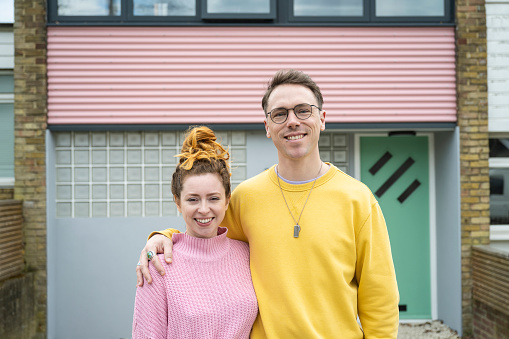 Waist-up view of Caucasian man and woman in casual clothing standing in front of modern style pastel two-story house and smiling at camera.