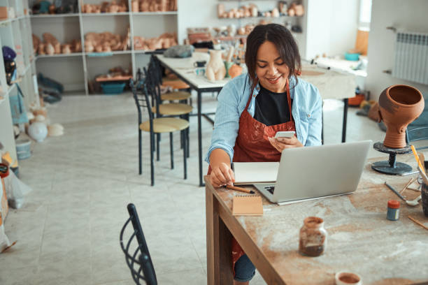istock Joyful woman using laptop and smartphone in pottery workshop 1319479722