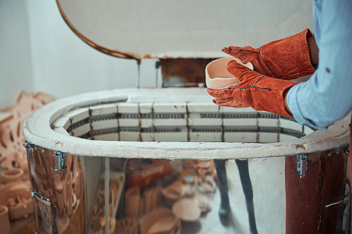 Close up of woman ceramist in gloves holding clay bowl while standing near electric ceramic kiln