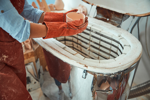 Close up of young woman ceramist holding handmade clay bowl while standing near electric ceramic kiln