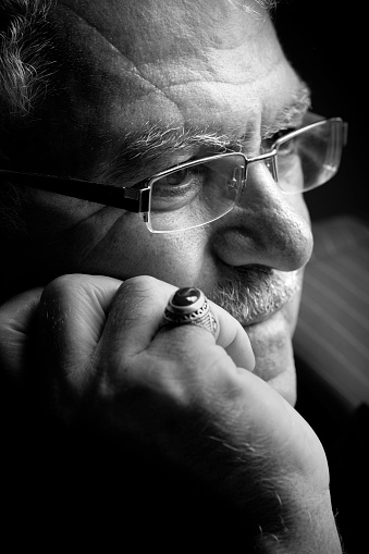 A portrait of 60-69 years old mature Turkish man. He has mustache and he puts his hands on his chin. He has a ring on his finger and he thinks some thinks. The photo was shoot indoors with daylight towards the black backgrounds.