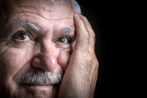 A portrait of 60-69 years old mature Turkish man. He has mustache and he puts his hand on his face. He thinks some thinks. The photo was shoot indoors with daylight towards the black backgrounds.