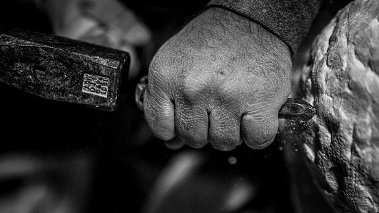 Close-up black and white photo of a stonemason's hand as he uses a hammer and chisel on a stone.