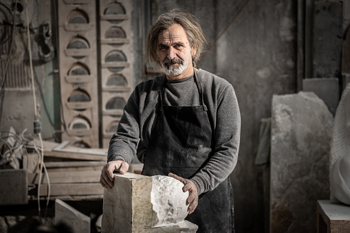 Portrait of a mature caucasian stone sculptor holding his work-in-progress sculpture and looking at the camera in his art studio.