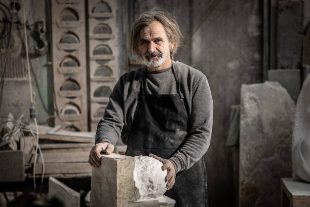 Portrait of a stone sculptor in his studio looking at the camera Portrait of a mature caucasian stone sculptor holding his work-in-progress sculpture and looking at the camera in his art studio. mason craftsperson stock pictures, royalty-free photos & images