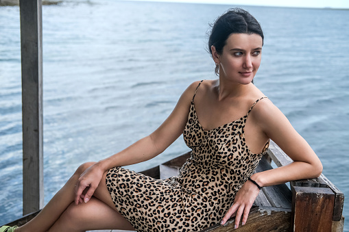 Young woman sitting by the lake or river during summer, wearing leopard pattern dress and enjoying the view