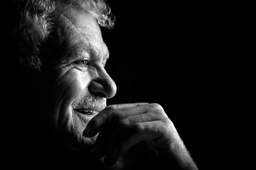 A portrait of 60-69 years old mature Turkish man. He has mustache and looking away. He's hand and faca are in the photo with black copy space. The photo was shoot indoors with daylight towards the black background.