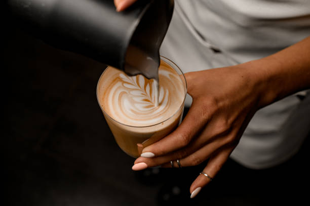 Close-up of woman barista gently drawing pattern pouring milk into glass with latte Close-up of woman barista gently drawing pattern pouring milk from jug into glass with latte in her hand milk froth stock pictures, royalty-free photos & images