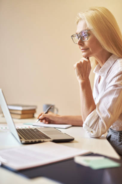 istock Focused mature businesswoman in glasses looking busy while using laptop, making notes, working in the office 1319490089
