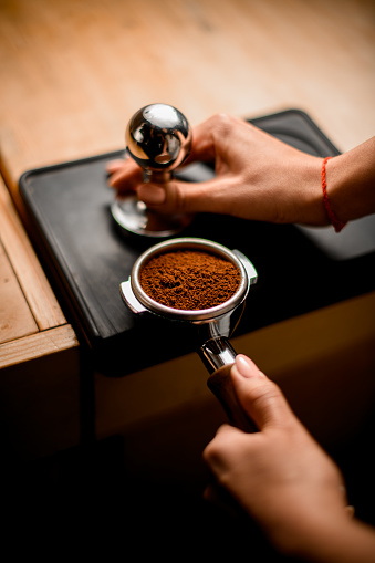 Close-up view of baristas hand tamping coffee in portafilter before making fresh drink in coffee machine at coffee shop.