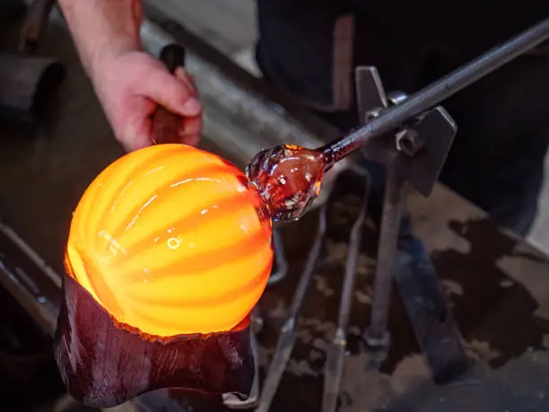 Glassblowing crafter rounds the hot-striped vase  with wet woode tool at the end of the glass blowing pipe. Traditional glass art workshop. Bohemia glass studio.