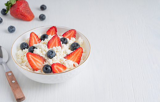 Quark fresh dairy product made of fermented soured milk also curd or cottage cheese served in bowl with spoon and fresh strawberries and blueberries on white wooden table. Image with copy space