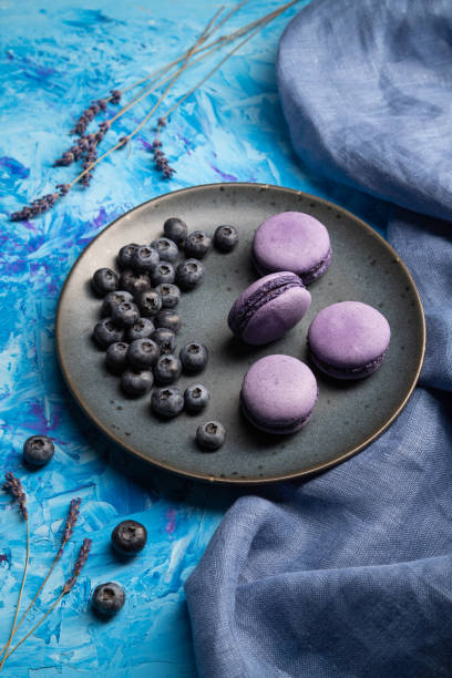 istock Purple macarons or macaroons cakes with blueberries on ceramic plate on a blue concrete background and blue textile. Side view, close up. 1319508328