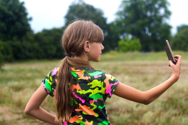 Defocus selfies. Little girl poses and takes a selfie by hand. Back view of Charming teen girl in summer standing on the meadow and taking a selfie on the phone. Nature background. Out of focus Defocus selfies. Little girl poses and takes a selfie by hand. Back view of Charming teen girl in summer standing on the meadow and taking a selfie on the phone. Nature background. Out of focus. child 10 11 years 8 9 years cheerful stock pictures, royalty-free photos & images