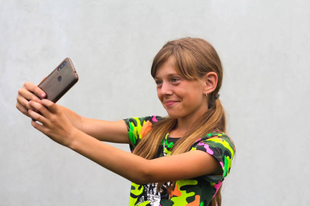 A ten year old girl poses and takes a selfie with her hand on her phone in a brown case. Gray white background. Smiling attractive girl is having fun at the camera in colorful clothes Selective focus A ten year old girl poses and takes a selfie with her hand on her phone in a brown case. Gray white background. Smiling attractive girl is having fun at the camera in colorful clothes Selective focus. child 10 11 years 8 9 years cheerful stock pictures, royalty-free photos & images