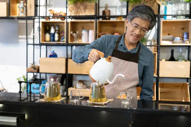 An older Asian waiter is pouring a cup of tea from a fancy teapot into a glass cup while he is standing behind the counter at a modern chinese tea shop