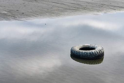 This is a full length photograph of a thrown away car tire washed up onshore of Crescent City beach in California, USA. The water on the shoreline reflects the clouds in the sky.