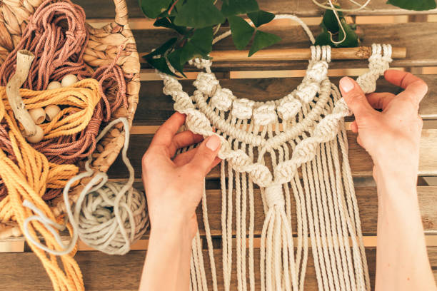 Close up of women's hands weaving macrame from white cotton threads in a home workshop.Home decor.Handmade concept. Close up of women's hands weaving macrame from white cotton threads in a home workshop.Home decor.Handmade concept.Selective focus with shallow depth of field,top view. macrame stock pictures, royalty-free photos & images