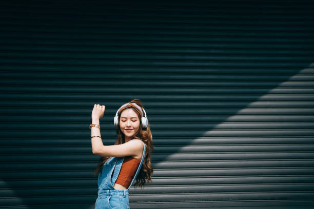 spensierata donna asiatica sorridente che balla con gli occhi chiusi mentre ascolta musica in cuffia all'aperto contro pareti colorate e luce solare. musica e stile di vita - fashionable foto e immagini stock