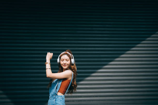 Carefree smiling young Asian woman dancing with her eyes closed while listening to music on headphones outdoors against coloured wall and sunlight. Music and lifestyle Carefree smiling young Asian woman dancing with her eyes closed while listening to music on headphones outdoors against coloured wall and sunlight. Music and lifestyle chinese woman stock pictures, royalty-free photos & images