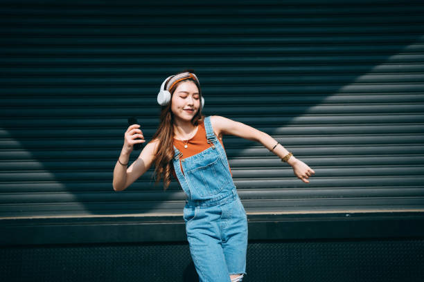 carefree smiling young asian woman holding smartphone, dancing with her eyes closed while listening to music on headphones outdoors against coloured wall and sunlight. music and lifestyle - youth and age imagens e fotografias de stock