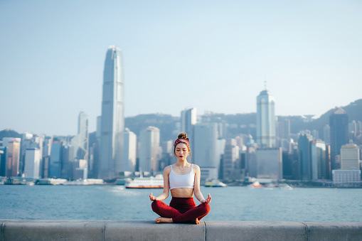 Young Asian sports woman meditating outdoors, practicing mindfulness yoga in the morning, against spectacular Hong Kong city skyline by the promenade of Victoria harbour. Girl power, wellbeing, fitness and healthy lifestyle concept