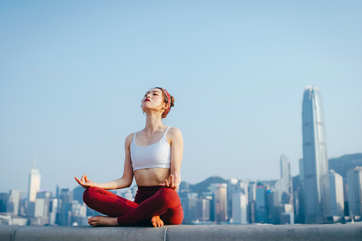 Young Asian sports woman meditating outdoors, practicing mindfulness yoga in the morning, against spectacular Hong Kong city skyline by the promenade of Victoria harbour. Girl power, wellbeing, fitness and healthy lifestyle concept